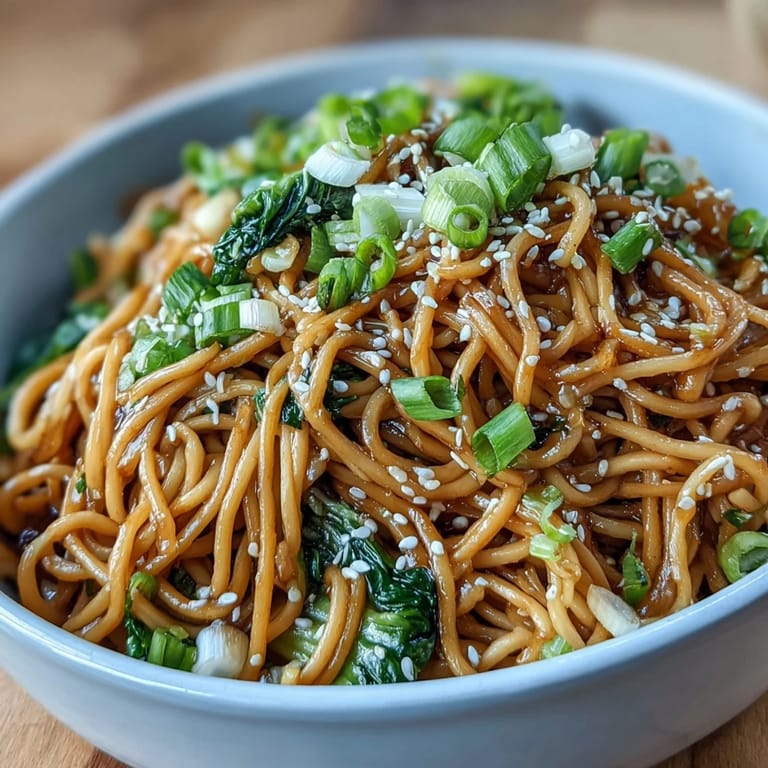 Close-up of Asian ginger-scallion noodles with sautéed greens, showcasing glossy sauce and colorful vegetable garnishes.